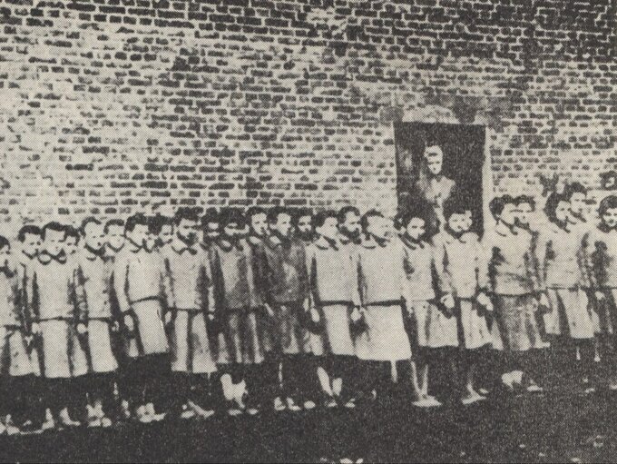 Dziewczęta na&nbsp;tle budynku podobozu dziewczęcego w&nbsp;obozie przy ul. Przemysłowej. W&nbsp;drzwiach stoi nadzorczyni Genowefa Pohl