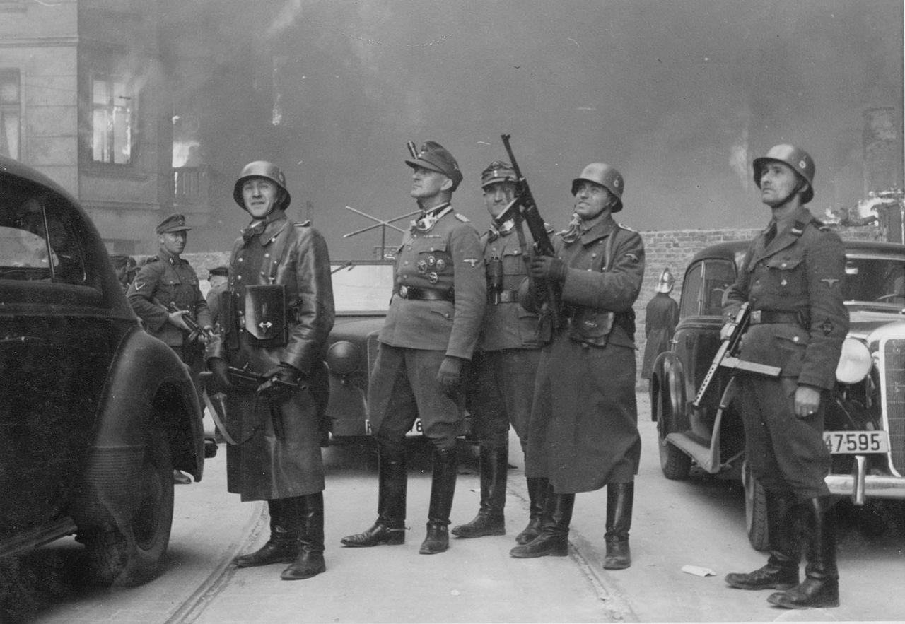 Fotografia z Raportu Stroopa. Oryginalny niemiecki podpis: "Dowódca wielkiej operacji". Generał SS Jürgen Stroop stoi pośrodku i patrzy do góry; z jego lewej strony Josef Blösche